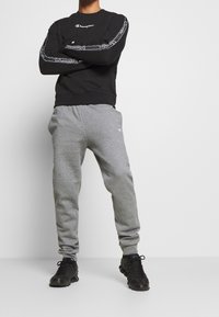 Felpa nera con logo e dettaglio bianco sui lati, abbinata a pantaloni grigi. Il modello indossa scarpe sportive nere. Outfit semplice e informale.