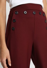 Pantalons bordeaux taille haute avec une texture lisse, présentant six boutons noirs sur le côté pour la fermeture et le détail.