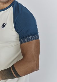 Heren overhemd met korte mouwen, met een marineblauwe bovenkant en een crèmekleurige onderkant. Voorzien van de tekst "SILK" op de mouw en een logo op de borst.