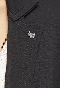 Svart tygblazer med en liten silverfärgad rosett-nåldekt och rhinestone-detalj på kragen, med rena sömmar och strukturerat tyg.