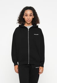 Felpa con zip nera, dotata di tasca frontale e polsini a costine. Presenta un logo argento sul lato sinistro del petto. Abbinata a pantaloni da jogging neri coordinati.