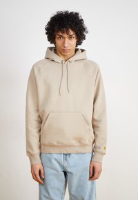 Beige hooded sweatshirt with a front pocket, ribbed cuffs, and hem, featuring a logo accent on the sleeve. Soft fabric, relaxed fit.