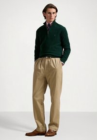 Green zip-neck sweater, beige pleated trousers, brown dress shoes, subtle patterns, relaxed fit, and woven texture.