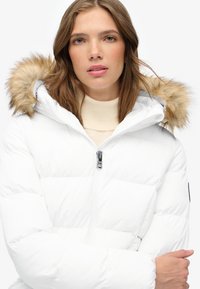 Chaqueta de plumón blanca con capucha forrada de piel, cierre de cremallera en la parte frontal, textura acolchada y detalles en hardware negro.