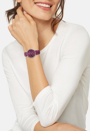 Montre à bracelet en cuir violet avec un boîtier rond en argent, des marqueurs blancs et argent, et un cadran violet foncé. Modèle portant un haut à manches longues blanc.