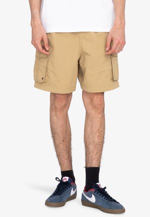 Beige cargo shorts with two side pockets, made from lightweight fabric. Styled with a white shirt and navy sneakers with red accents.