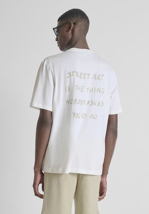 Uomo con occhiali neri, maglietta bianca con scritta beige "Street art is the thing nobody asked you to do" e pantaloncini beige con texture, di spalle.