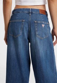 Jeans in denim con vestibilità rilassata, colore blu, cinque tasche e una toppa con logo a triangolo sulla tasca posteriore destra. Dettaglio in pelle sulla cintura.
