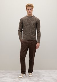 Bruine gebreide trui met een gestructureerd patroon, gecombineerd met bruine broeken en witte sneakers met beige accenten, staand tegen een eenvoudige achtergrond.