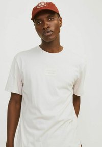 Tricou alb din bumbac, cu guler rotund și mâneci scurte, având un patch rectangular cu logo. Asortat cu o șapcă maro.