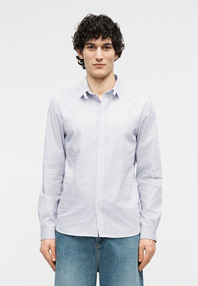 Νεαρός με κοντά σγουρά σκούρα μαλλιά, ντυμένος με ανοιχτό μπλε πουκάμισο με κάθετες ρίγες και μπλε τζιν, στέκεται μπροστά σε ένα απλό φόντο.