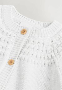 Bela pletena kardiganj s teksturiranim jočnim delom, rebrastim ovratnikom in dvema lesenima gumboma spredaj. Mehka, lahka tkanina.