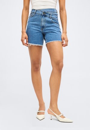 Kobieta w niebieskich szortach dżinsowych z wysokim stanem i surowym wykończeniem oraz białych, spiczastych butach na obcasie typu slingback, stojąca na jednolitym tle.