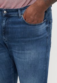 Mano di pelle scura parzialmente dentro la tasca anteriore di jeans denim blu medio con zone sbiadite, indossando una maglietta rossa.