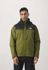 Giacca impermeabile verde con pannello nero sul petto, cappuccio regolabile e zip frontale. Presenta il logo sul lato sinistro del petto e una texture morbida.