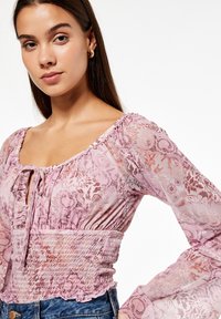 Jasno różowa bluzka w kwiaty z przezroczystymi rękawami, kwadratowym dekoltem i marszczonym pasem. Posiada falbany na brzegach oraz skomplikowane wzory kwiatowe.