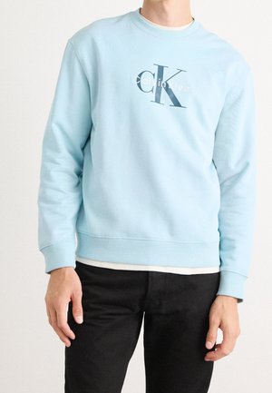 Mand iført lyseblå Calvin Klein sweatshirt over hvid skjorte, kombineret med sorte bukser, stående mod en ensfarvet baggrund.