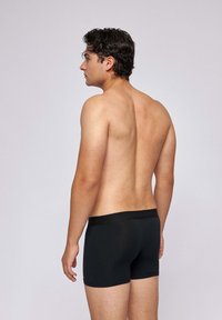 Male model with dark hair stands facing left, shirtless, wearing black boxer briefs against a plain light gray background.