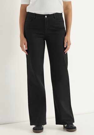 Vrouw die zwarte broek met wijde pijpen, wit topje en zwarte sneakers draagt, staand tegen een effen lichte achtergrond.