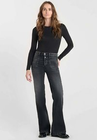 Top negro de manga larga combinado con jeans de mezclilla oscura de talle alto y corte acampanado, con detalles desgastados y cierre de doble botón.