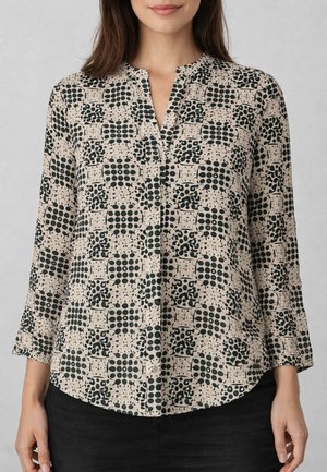 Donna che indossa una camicetta beige con motivi geometrici e astratti neri, caratterizzata da una scollatura a V e maniche lunghe, abbinata a pantaloni neri.