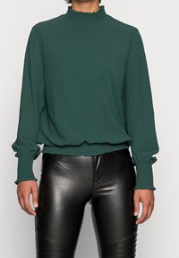 Grön långärmad blus med fransig hög krage och rynkade ärmslut. Buren över svarta fuskskinnbyxor. Slät textur.