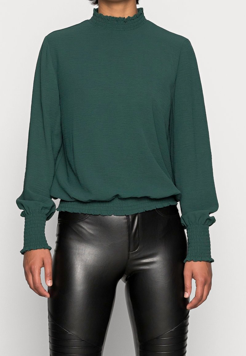 Grön långärmad blus med fransig hög krage och rynkade ärmslut. Buren över svarta fuskskinnbyxor. Slät textur.