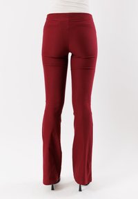 Pantaloni rossi, aderenti, elasticizzati con orlo svasato, texture liscia, vita media e dettagli di cucitura minimi. Posizionati su uno sfondo chiaro.