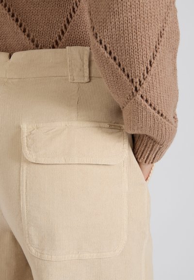 Beige corduroy broek met een zak op de achterkant, gecombineerd met een gemêleerd bruin gebreid trui, die de ribbeltextuur van de stof benadrukt.