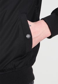 Giubbotto bomber nero con una texture liscia, dotato di polsini a coste e un dettaglio con logo circolare sulla manica. Inquadratura ravvicinata della mano in tasca.