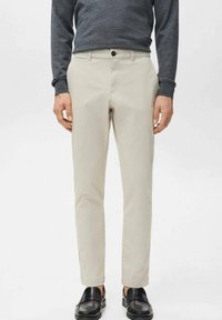 Mężczyzna ubrany w beżowe wąskie spodnie, ciemną koszul�ę z długim rękawem i czarne mokasyny, stojący na tle jednolitego białego tła.