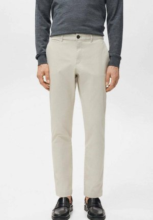 Mężczyzna ubrany w beżowe wąskie spodnie, ciemną koszulę z długim rękawem i czarne mokasyny, stojący na tle jednolitego białego tła.