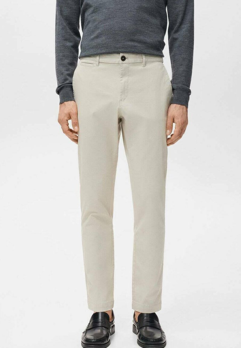 Mężczyzna ubrany w beżowe wąskie spodnie, ciemną koszulę z długim rękawem i czarne mokasyny, stojący na tle jednolitego białego tła.