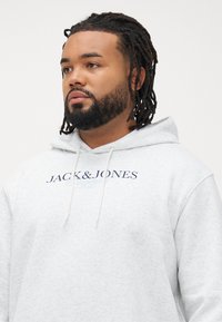 Mann med flettet hår og skjegg iført en lys grå Jack & Jones hettegenser, ser litt til venstre mot en ensfarget bakgrunn.
