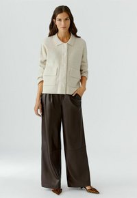 Crème-kleurige gebreide cardigan met een kraag, knoopsluiting en twee voorzakken, gecombineerd met donkerbruine leren broek met wijde pijpen.