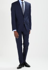 Marineblauw pak met een enkele knoopsluiting, notch-revers en twee voorzakken. Gecombineerd met een lichtblauw overhemd en een grijze stropdas, zwarte schoenen.