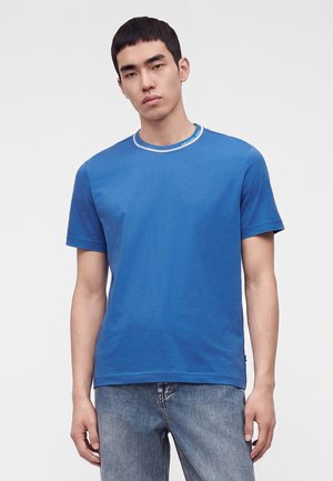 Jeune homme portant un t-shirt bleu uni avec un liseré blanc au col et un jean bleu délavé, debout devant un fond blanc uni.