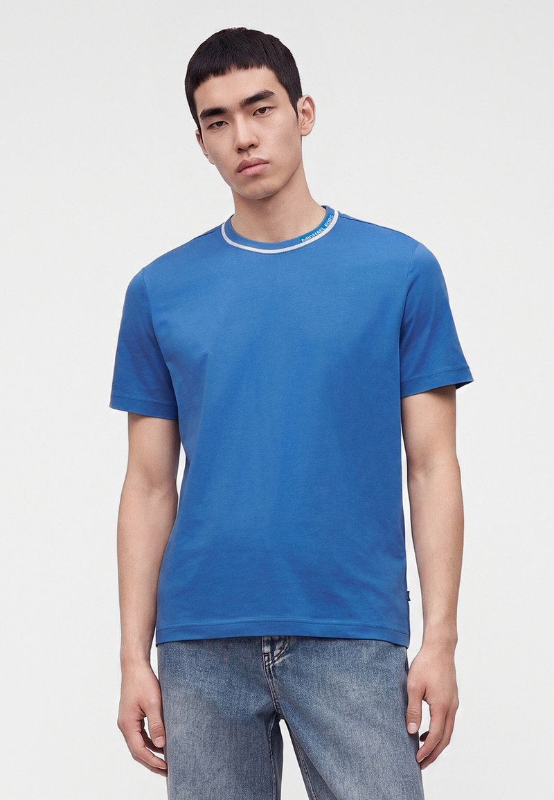 Jeune homme portant un t-shirt bleu uni avec un liseré blanc au col et un jean bleu délavé, debout devant un fond blanc uni.