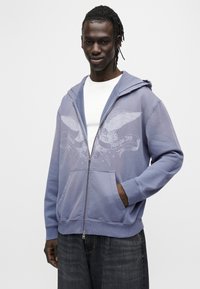 Μωβ φούτερ με φερμουάρ από απαλό υλικό, με γραφικό τύπωμα φτερών και το κείμενο "Analogue Days". Τσέπες καγκουρό και κουκούλα με κορδόνι.