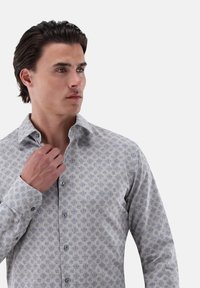 Maglietta grigia a maniche lunghe con un sottile motivo geometrico blu. Presenta un colletto classico, chiusura con bottoni e polsini. Tessuto morbido.