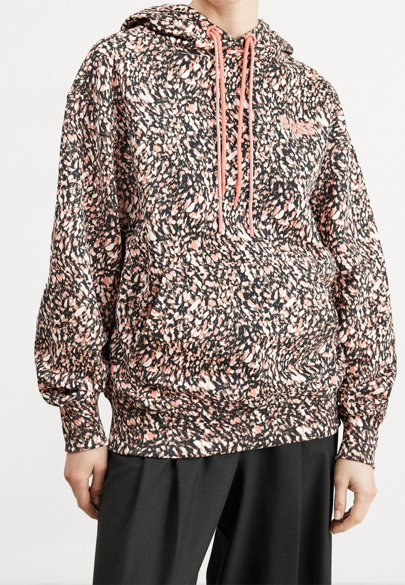 Personne portant un sweat à capuche ample et à motifs avec des taches abstraites roses et noires et des cordons roses, associé à un pantalon noir.
