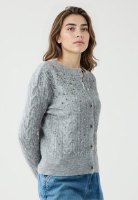 Grijze gebreide cardigan met een gestructureerd kabelontwerp en decoratieve versieringen. Het heeft een ronde halslijn en knopen aan de voorkant.