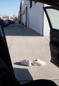 Baskets blanches et roses placées côte à côte sur un trottoir ensoleillé vues à travers une porte de voiture ouverte dans une rue urbaine calme.
