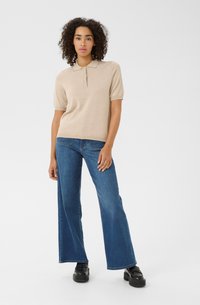 Vrouw staat in een beige polo met korte mouwen, brede blauwe jeans en zwarte platform schoenen tegen een effen witte achtergrond.