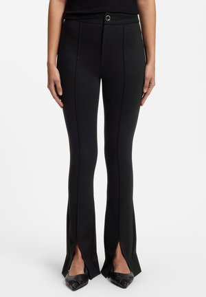 Pantaloni neri aderenti con dettaglio della cucitura frontale e spacchi anteriori alle caviglie, indossati con scarpe nere a punta, mostrati dalla vita ai piedi.