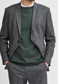Szary, strukturalny żakiet na tle ciemnozielonego swetra, z delikatnym wzorem i dopasowanym krojem. Całość uzupełnia biała kołnierzyk koszuli.