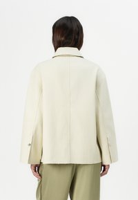 Cream-colored jacket with a relaxed fit, wide collar, and buttoned cuffs. Made from smooth fabric, features a center seam at the back.