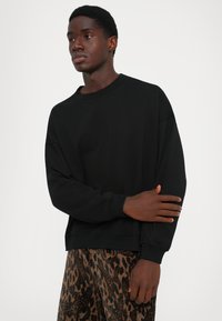 Jeune homme à la peau foncée portant une chemise noire à manches longues et un pantalon marron à motifs, ayant une expression contemplative avec les bras croisés.