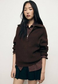 Maglione a coste marrone con collare a mezza zip, indossato sopra una camicia con colletto marrone scuro, abbinato a una gonna nera plissettata.