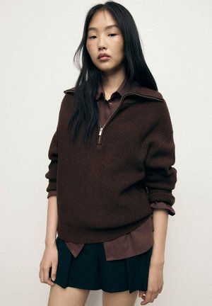 Pull en tricot côtelé marron avec col zippé mi-haut, superposé à une chemise à col marron foncé, associé à une jupe plissée noire.